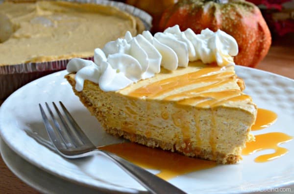 A slice of pumpkin cheesecake on a graham cracker crust with whipping cream and caramel drizzled on top.