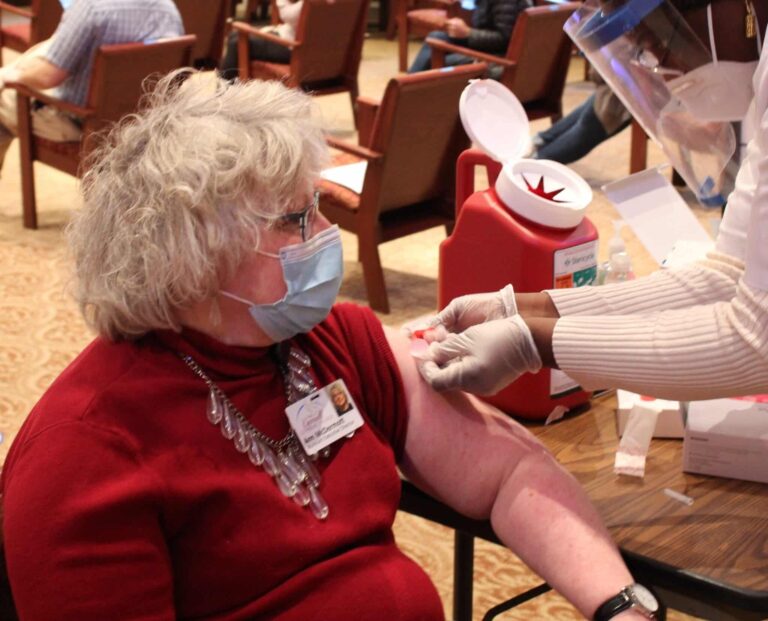 Assistant executive director, Ann McDermott, sitting in chair getting a band-aid placed on her arm after receiving the COVID-19 vaccine.