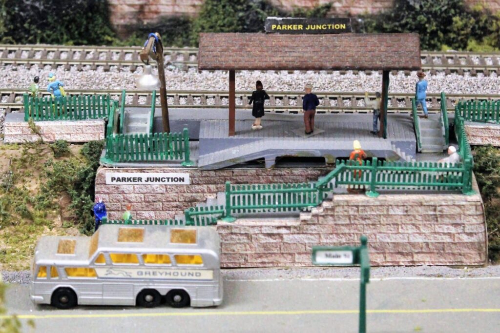 HO model railroad of Parker Junction with models of a Greyhound bus driving by and people waiting on the platform for the train.
