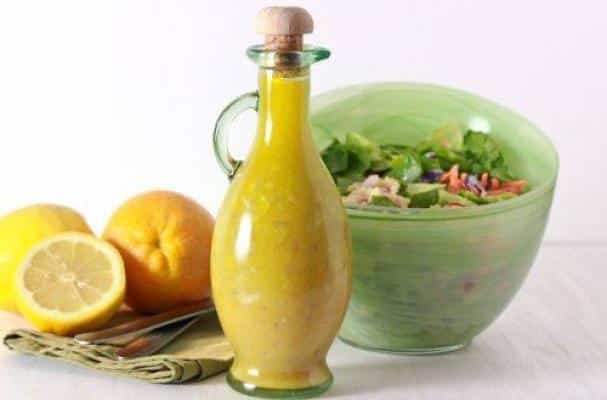 A bottle of citrus vinaigrette in front of lemons, an orange, and a salad bowl.