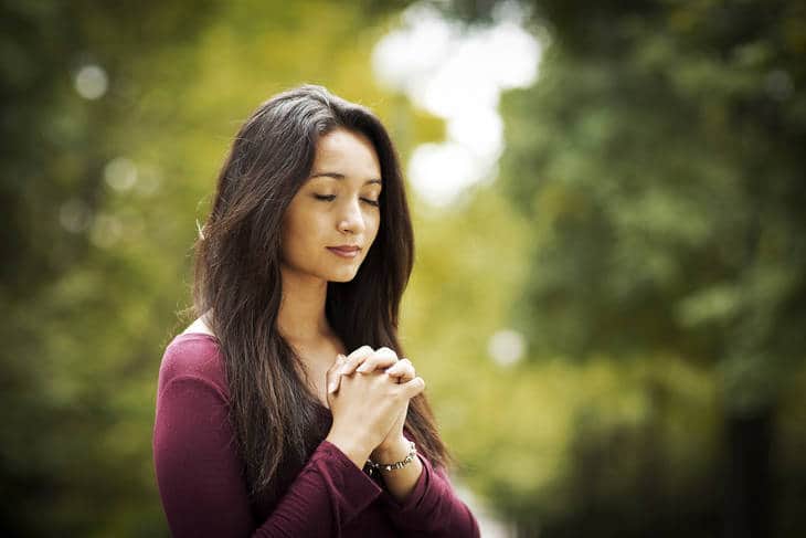 A woman with her clasped hands at her heart and her eyes closed enjoying the peace of being surrounded by trees.