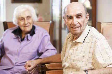 A caregiver husband is holding hands with his wife seated in chairs.