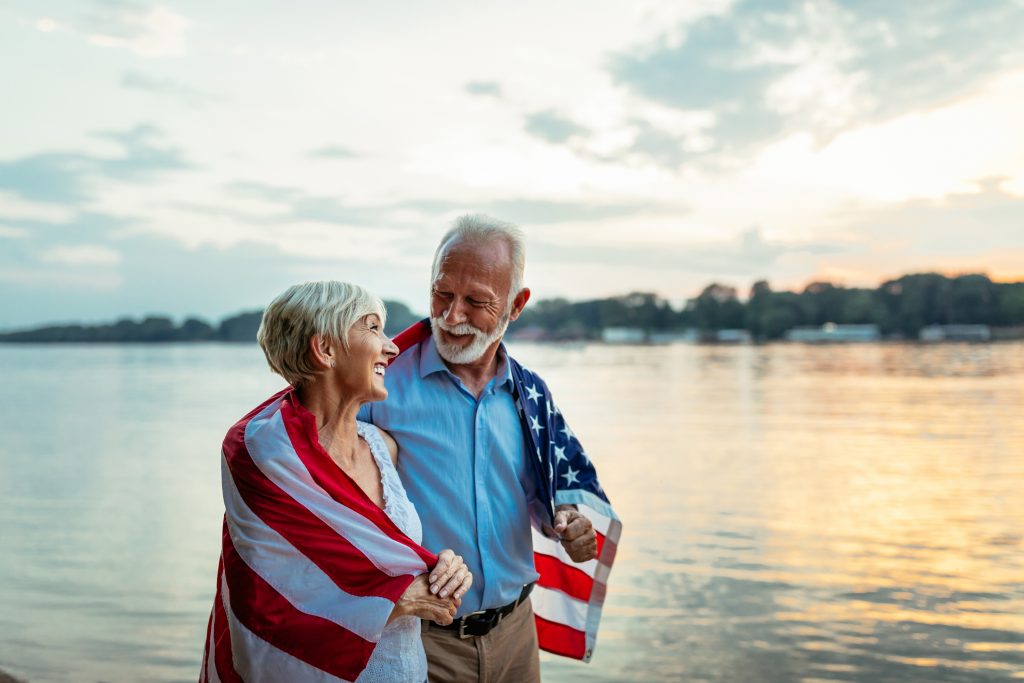 Senior veteran and spouse, draped in the American flag, are smiling because the VA's Aid and Attendance benefit is helping them with their senior living costs.