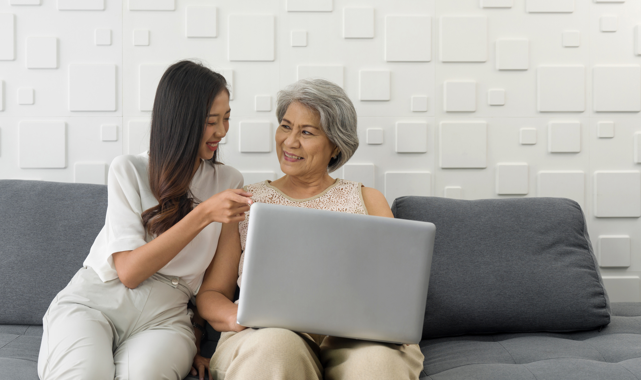 A young Asian woman sitting with her Mom, on a couch, looking at a computer and discussing if senior living is right for her Mom.