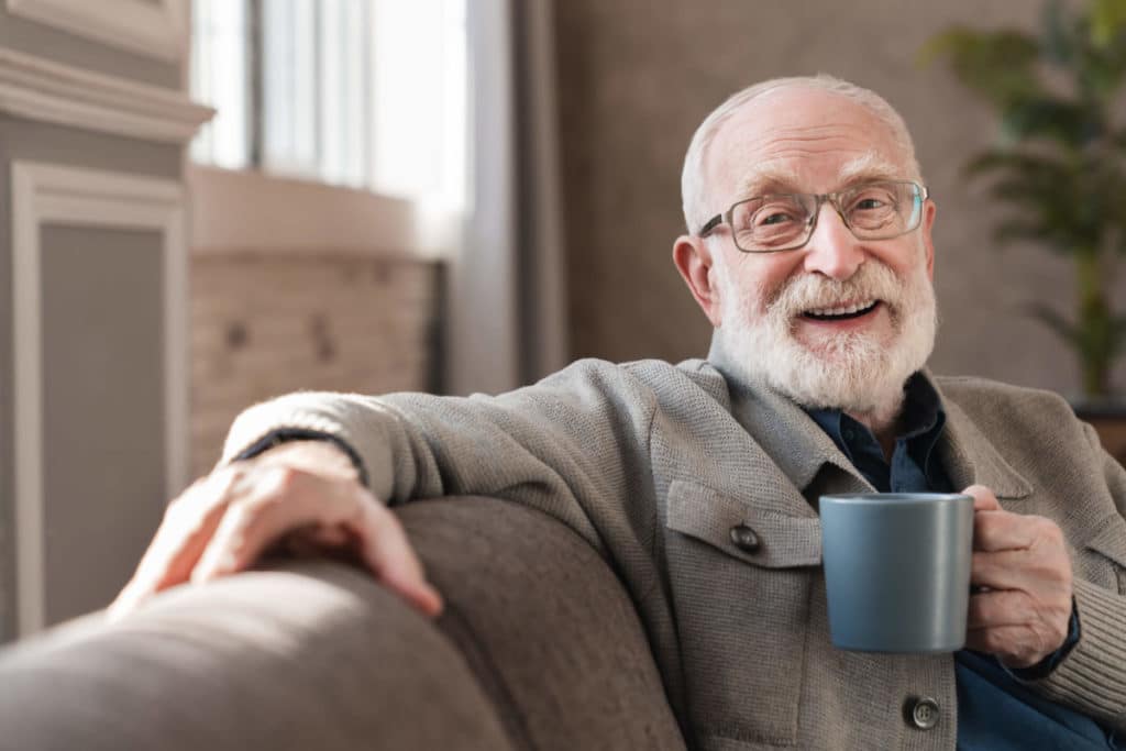 Senior male sitting on couch drinking out of a mug, smiling for having followed tips for deciding if senior living lifestyle is right for you