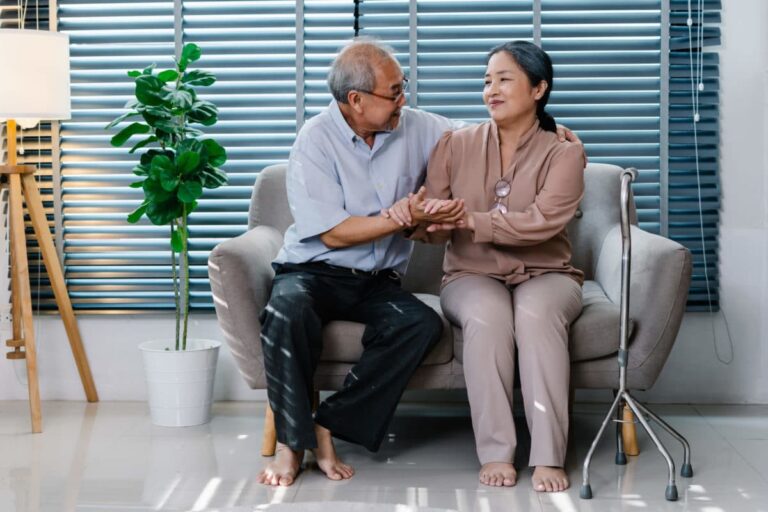 A husband provides at-home care by helping his wife from a couch.