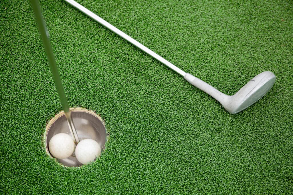 Putting golf club on green grass with golf ball in the hole - top view