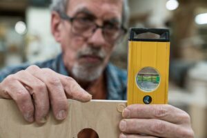 A senior man with glasses and beard in a woodworkers shop, using a spirit level checking his work.