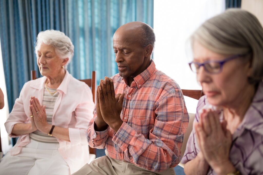 Senior man amidst women praying while sitting on chairs